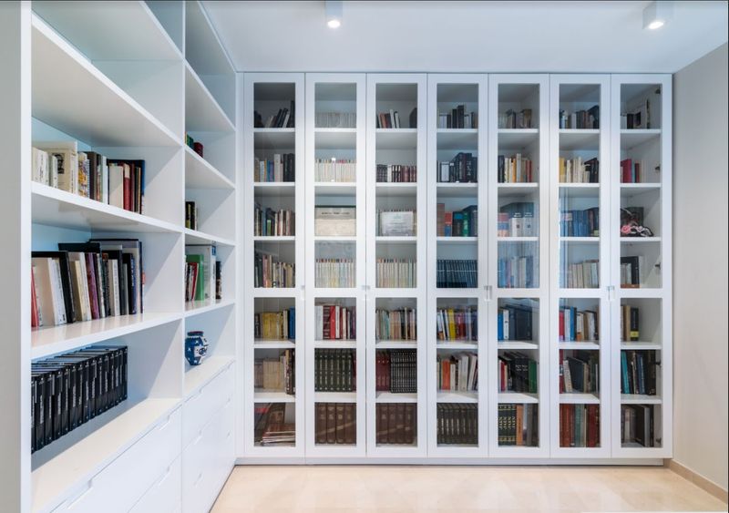 Practica-y-bonita-libreria-realizada-a-medida-lacada-en-blanco-con-puertas-de-cristal-enmarcadas-en-madera-lacada-blanca-min